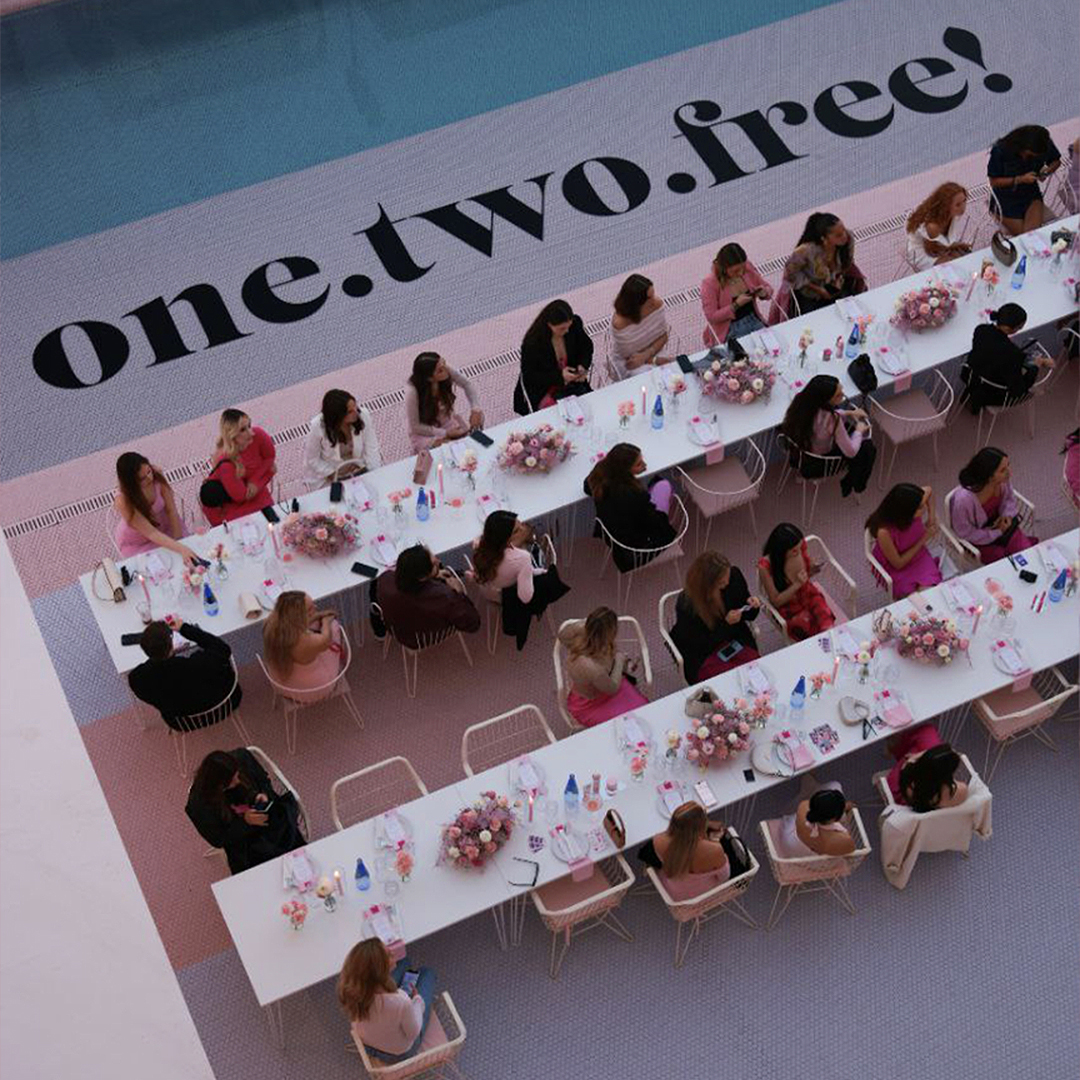 Two long tables near a pool with several woman sitting on the table. The pool is filled with water and on the ground is written one.two.free!